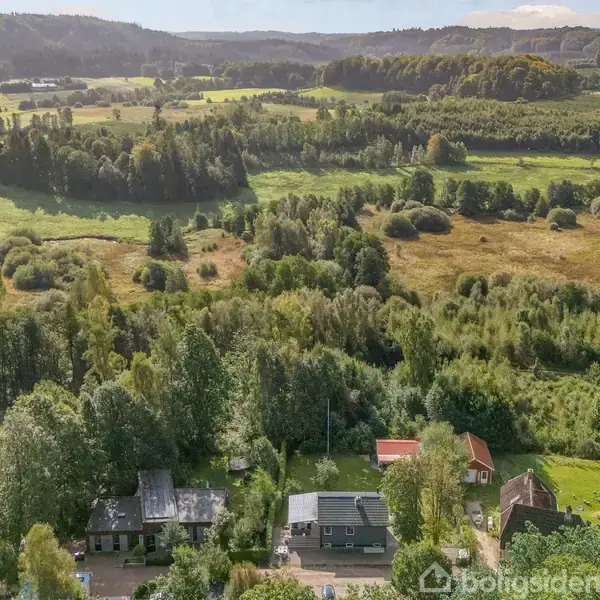 Huse ligger blandt træer i en grøn, bakket landskabskontekst med skove og åbne marker. I baggrunden ses bølgende bakker under en klar himmel.