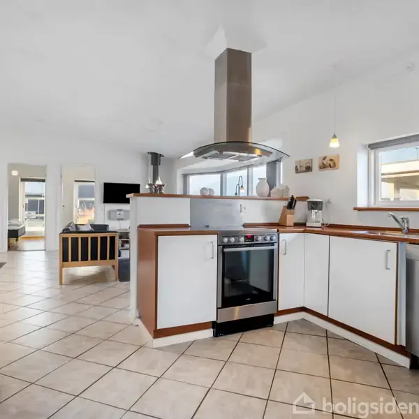 Et moderne køkken med en central kogeø og ståloverflader. Køkkenet har hvide skabe, en opvaskemaskine og flisegulv. I baggrunden ses en stue med sofa og tv.