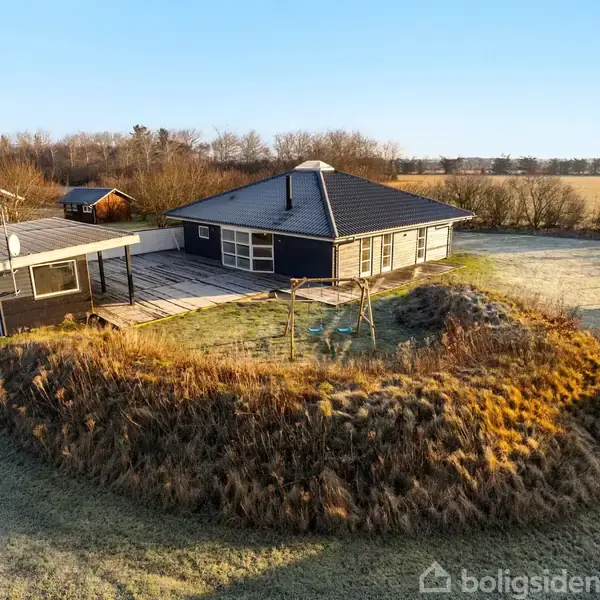 Et enkelt sort-hvidt sommerhus med en stor terrasse står i en græsgrøn have omkranset af buske og et hegn. Andre små huse ses i baggrunden på en solrig dag.