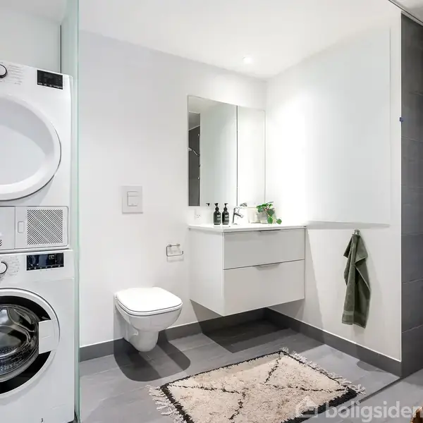 Vaskemaskine og tørretumbler står stablet i et moderne badeværelse. En væghængt håndvask med spejl og opbevaring er ved siden af toilettet. Håndklæde hænger på væggen. En tæppe ligger på gulvet.