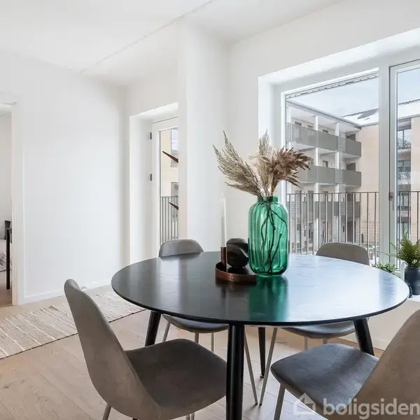 Rundt spisebord med fire stole, dekoreret med grøn vase og tørrede blomster, står i lyst rum med store vinduer. Til venstre ses en åben dør til et soveværelse.