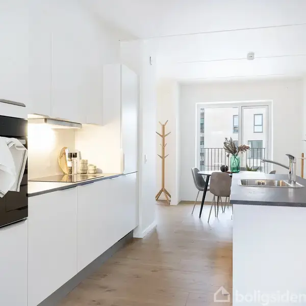 Et moderne køkkenområde med hvide skabe og en indbygget ovn, håndklæde hængende udenpå. I baggrunden ses en spiseplads med træbord og stole samt udsigt til en altan.