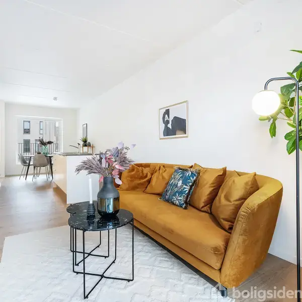 Blød, sennepsfarvet sofa står mod en hvid væg med billedkunst, blomstervase på sort bord foran, stor plante ved siden af, moderne, lyst rum med trægulv og spisestue i baggrunden.
