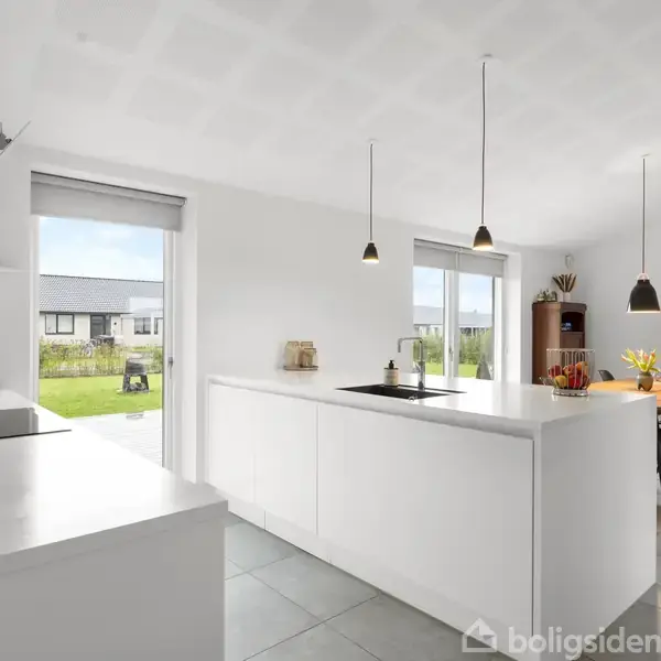 Lyst køkken med hvidt køkkenbord, kogeplader og vask. Tre sorte pendellamper hænger over bordet. I baggrunden ses spisebord med blomster og udsigt til en solrig have.