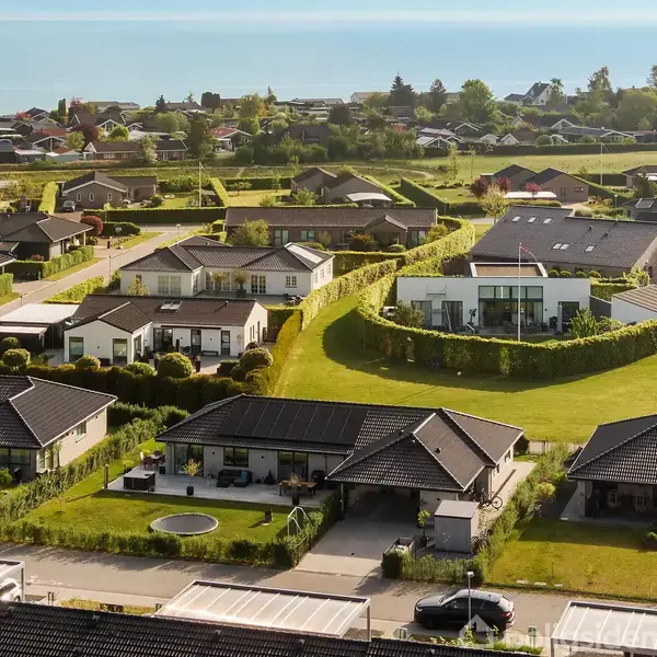 Huse ligger stille i et landområde med velplejede græsplæner og hække. I baggrunden er der udsigt til en fjern kystlinje og klar blå himmel.