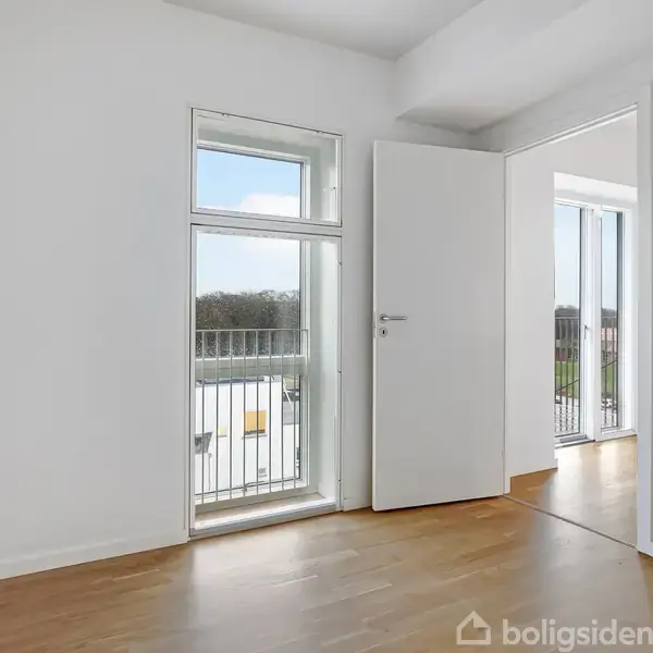 Et lyst rum med trægulve; en dør står åben mod en tilstødende balkon med glasrækværk og udsigt til træer. Hvidmalede vægge skaber en ren og moderne atmosfære.