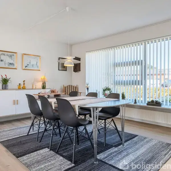Spisebord med seks sorte stole står på et stort tæppe. Ved siden af er en skænk med planter og billeder. Store vinduer med persienner giver udsigt til en terrasse.
