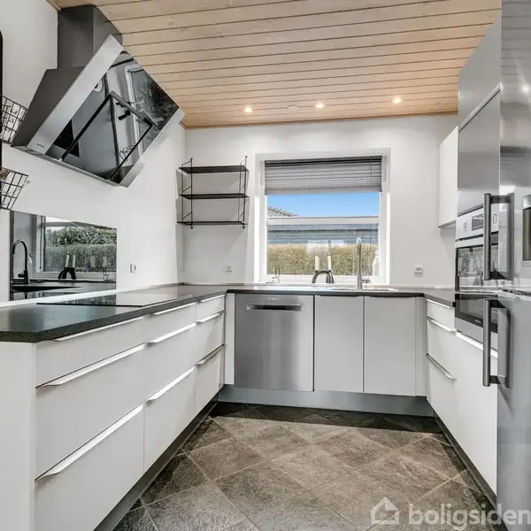 Et moderne køkken med hvide skabe, en stål-opvaskemaskine og køleskab, placeret i et lyst rum med trægulv og loft, vindue med udsigt til en grøn have.