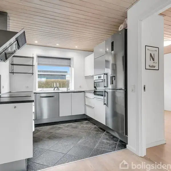 Et moderne køkken har hvide skabe og stålapparater. En plakat på væggen siger "Provence N Noïr". Køkkenet har flisegulv og et vindue med udsigt til en grøn have.