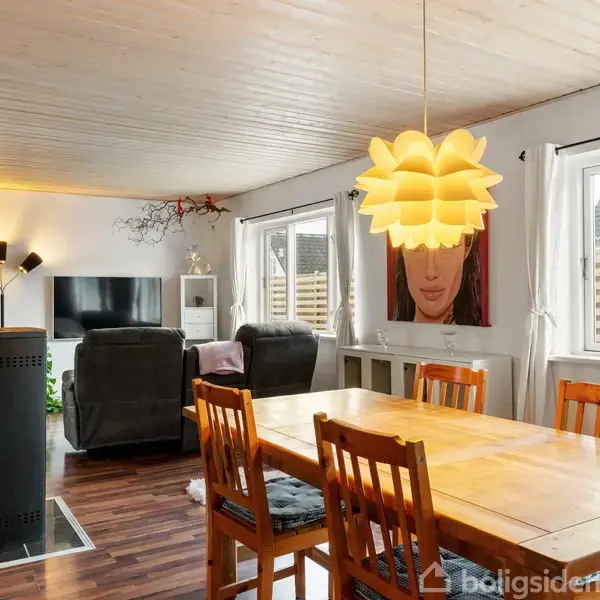 En spisebord med træstole står i en stue med en moderne lampe hængende over bordet. I baggrunden ses to lænestole, et TV og kunst på væggen.