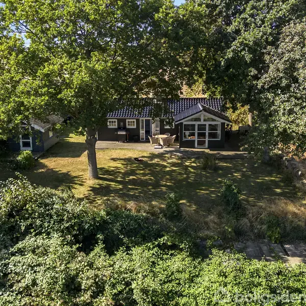 Hus med sort tag står i en solrig have omgivet af store træer. En terrasse med havemøbler ses foran huset, mens et skovkantsområde omgiver scenen.