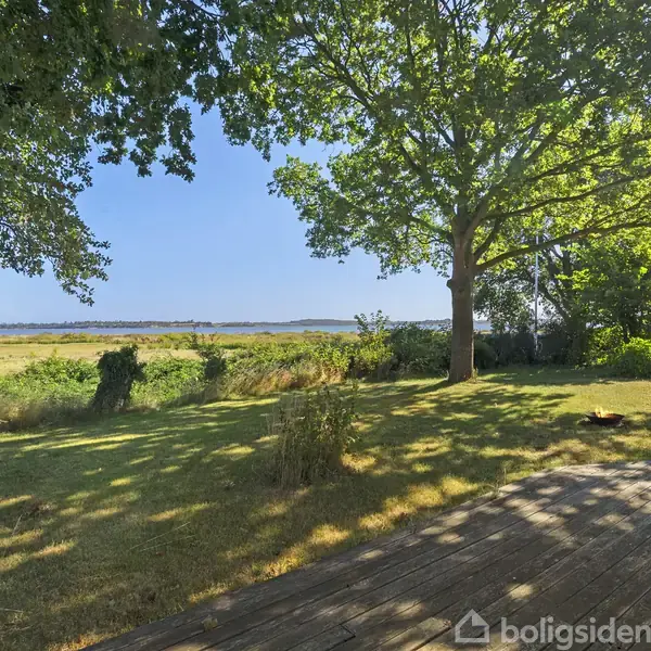 En træterrasse grænser op til en græsplæne med spredte træer. I baggrunden ses en udsigt over en eng med skove og himmel.