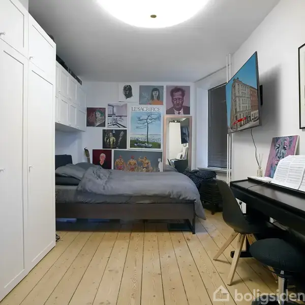 Et soveværelse med en seng dækket af gråt sengetøj står foran en væg dekoreret med plakater. Et klaver ved siden af har et åbent nodehæfte, og en stor fladskærm hænger på væggen.