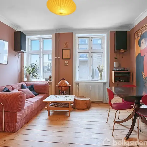 En rød sofa står langs en væg, pyntet med puder i et stueområde. Et bord står foran sofaen. Rummet har lyse vægge, trægulv og billeder på væggen.