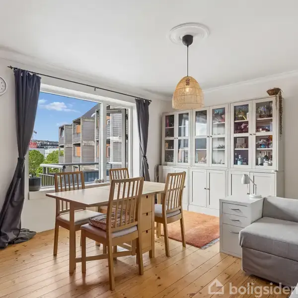 Spisestue med spisebord og stole, åben mod en balkon. En grå sofa står mod højre, og et vitrineskab mod bagvæggen. Røde og hvide vægge, vægur. Lysindfald fra venstre.