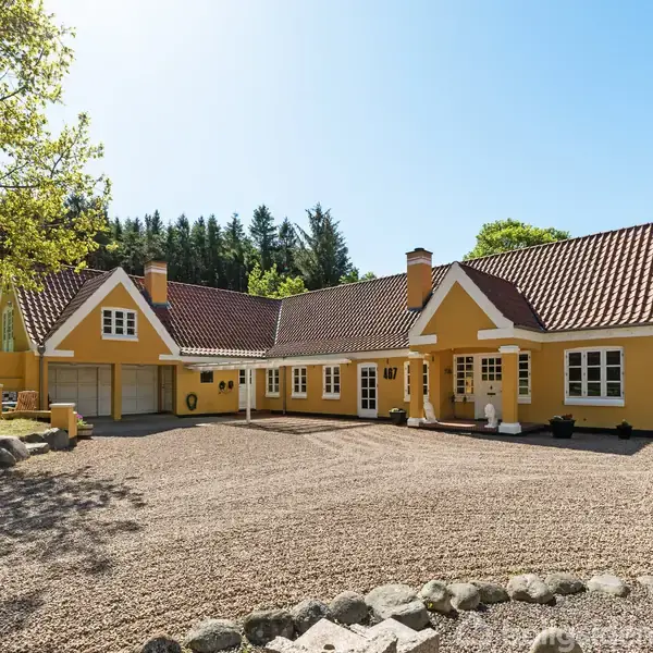 Gult hus med hvidt træværk og rødt tegltag står på en grusbelagt gårdsplads omgivet af grønne træer. Sollys skinner klart, hvilket giver en lys og indbydende atmosfære.