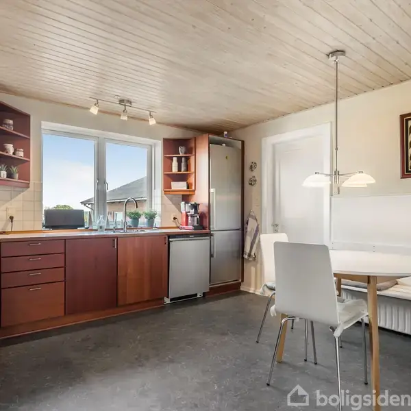 Et køkken med mørke træskabe og en hvid bordplade. To stålvaske er placeret under et stort vindue. Der er et rundt spisebord med stol og bænk i hjørnet.