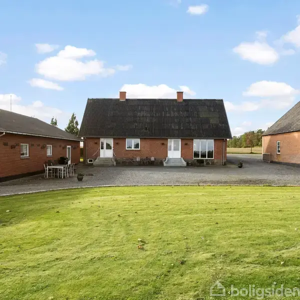 Et rødt murstenshus står stille mellem to sidebygninger, omgivet af en grusvej og frodig græsplæne under en blå himmel med spredte skyer.