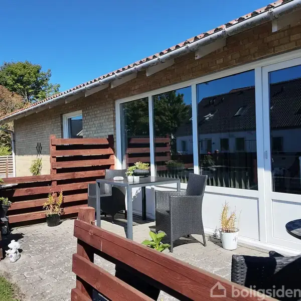 Hus med terrasse møbleret med bord og stole, omgivet af træhegn. Klare vinduer reflekterer en solrig himmel. Plantekrukker dekorerer området, placeret i en grøn have med træer i baggrunden.