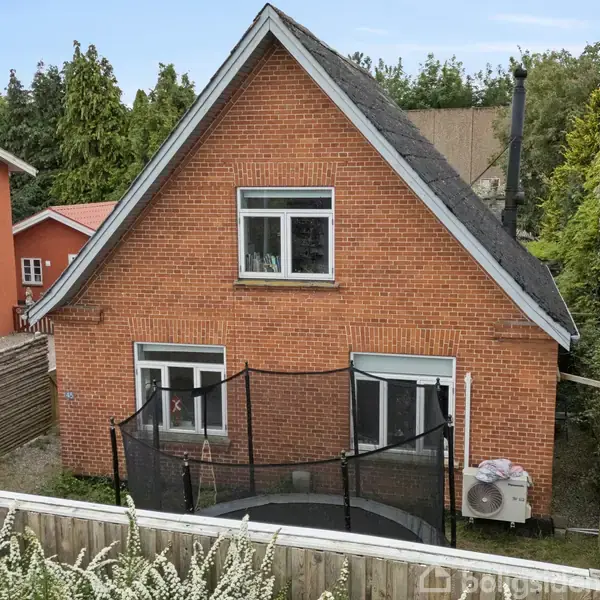 Et rødt murstenshus med skråt tag står i en have omgivet af hegn og træer. Foran huset er der en trampolin med sikkerhedsnet. To parkerede biler ses til venstre.