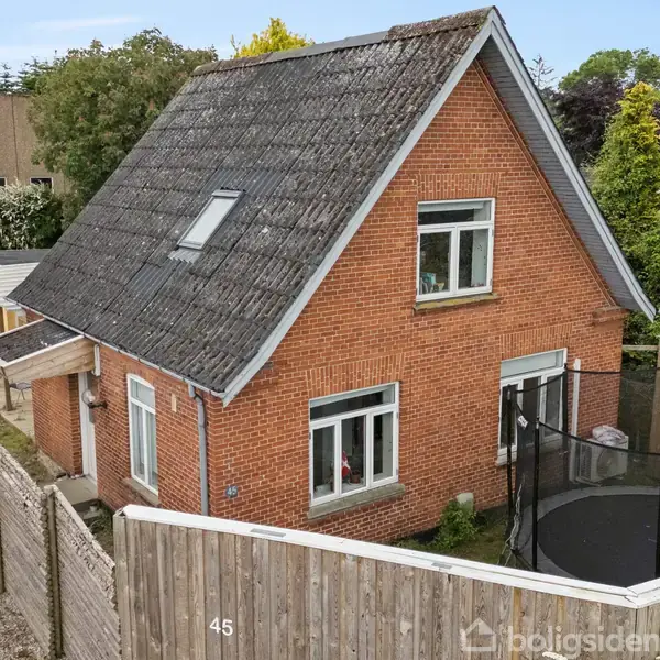 Et rødt murstenshus med skråtag har et hegn med nummer 45, en trampolin i haven og er omgivet af træer.