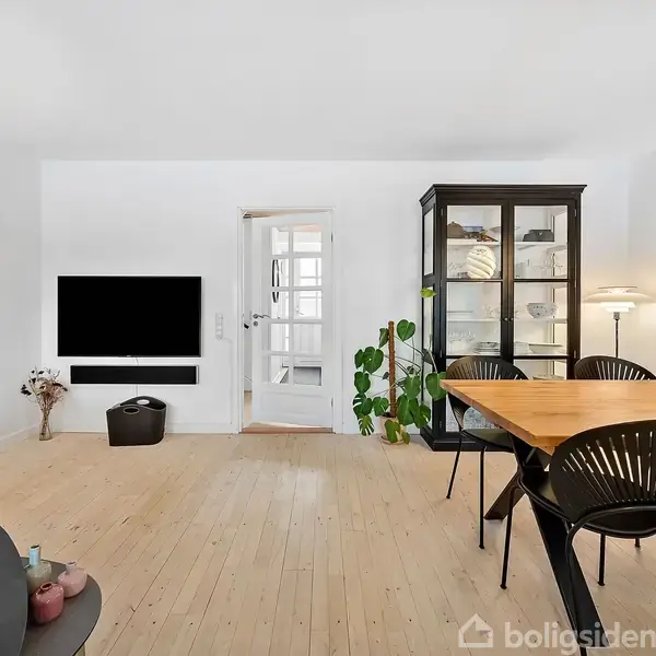 Stue med trægulv, stort spisebord og sorte stole i forgrunden; reol, lampe og TV på væggen bagved; dør i baggrunden. Indretningen er moderne og minimalistisk.