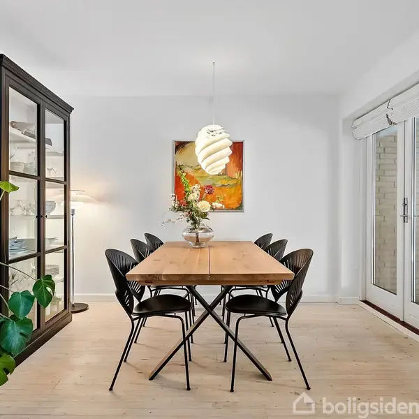 Et spisebord med seks sorte stole står under en moderne lampe. Ved siden af er et vitrineskab. Væggene er hvide, og der er en farverig maleri bagved.