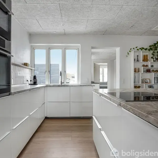 Moderne køkken med hvide skabe og gråt marmorbordplade, elektriske ovne indbygget i væggen. Rummet har lyse trægulve, stort vindue i baggrunden og en hylde med planter til højre.