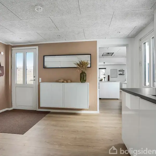 Vaskemaskine står i en lys gang med lysebrunt gulv og hvidmalede vægge. En dør leder udenfor, mens en skabsenhed med vase og dekorationer står ved siden af.