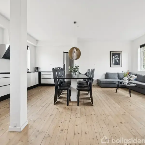 Et lysfyldt rum med et sort spisebord og stole i centrum, omgivet af et åbent køkken til venstre og en grå sofa med et sofabord til højre. Billeder hænger på væggene.