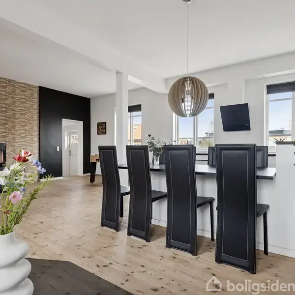 Rundt spisebord med sorte stole står i et lyst rum. En moderne lampe hænger over bordet, mens friske blomster i en vase pynter. Pejs ses i baggrunden.