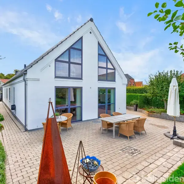 En moderne, hvidmalet hus med store glaspartier står omgivet af en velplejet have. Terrassen har et spisebord og en parasol, ved siden af en græsplæne og træer.