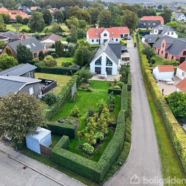 Et moderne hvidt hus står i en række af andre huse langs en smal vej. Huset har en grøn have med planter omkranset af hække i et boligområde.