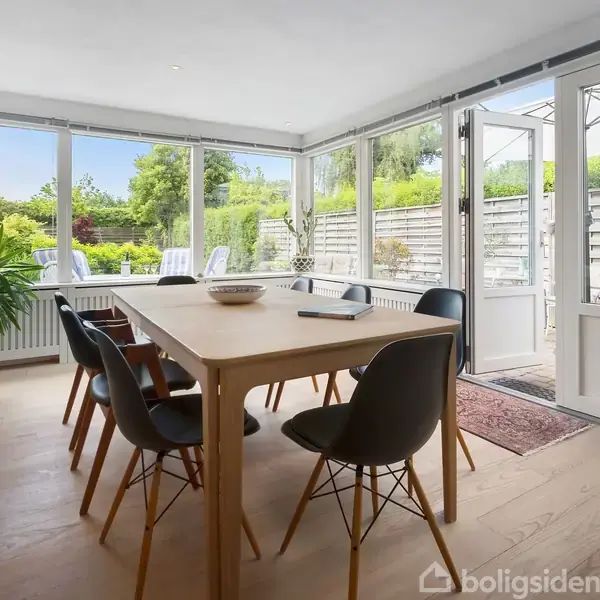 Seng med hvidt sengetæppe står stille ved store vinduer, der viser udsigt over frodige træer udenfor. Et maleri hænger på væggen ved siden.