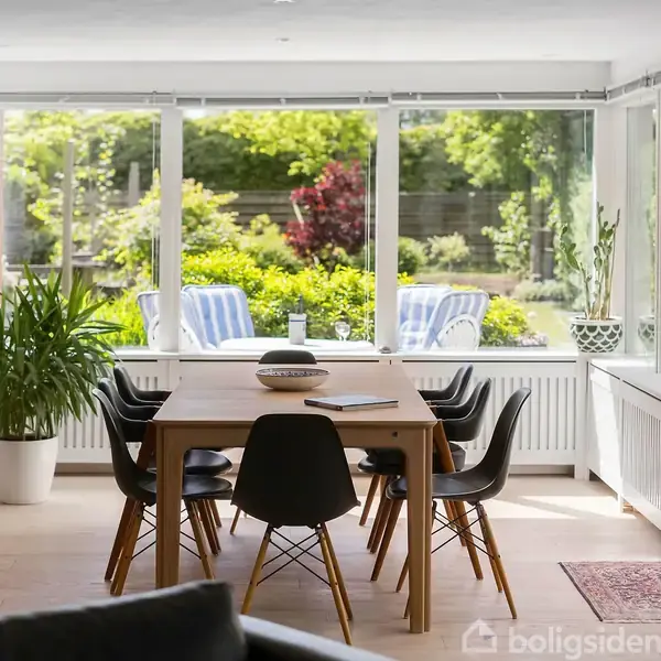 Et spisebord omgivet af seks sorte stole står i et rummeligt, lyst værelse med trægulv. Store vinduer og en åben dør viser ud til en terrasse med stole og grøn beplantning udenfor.