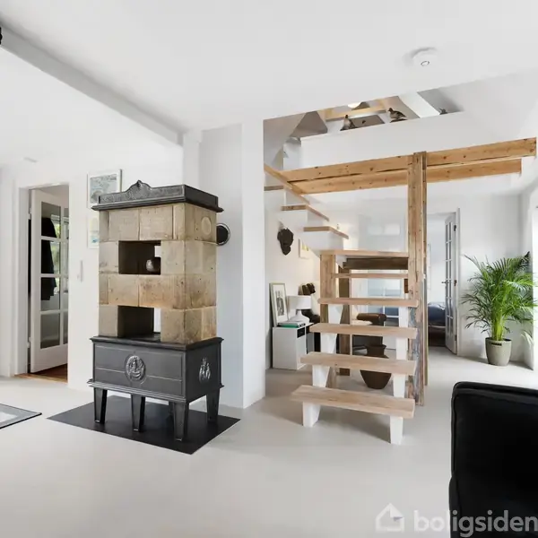 Hvidt hus med sort tegltag og store vinduer i udestue. Beliggende i en gårdhave med brostensbelægning, omgivet af planter og en grøn hæk.