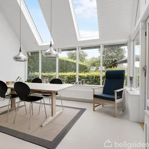 Et lyst spiseområde indeholder et bord med fire sorte stole, oplyst af to loftslamper. Store vinduer og ovenlysvinduer giver udsigt til en grøn have.