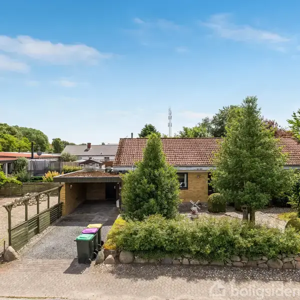 Et gulmurstenshus med rødt tegltag er omgivet af en velplejet have med træer og buske. En carport med skraldespande står ved indkørslen. Baggrunden har flere huse.