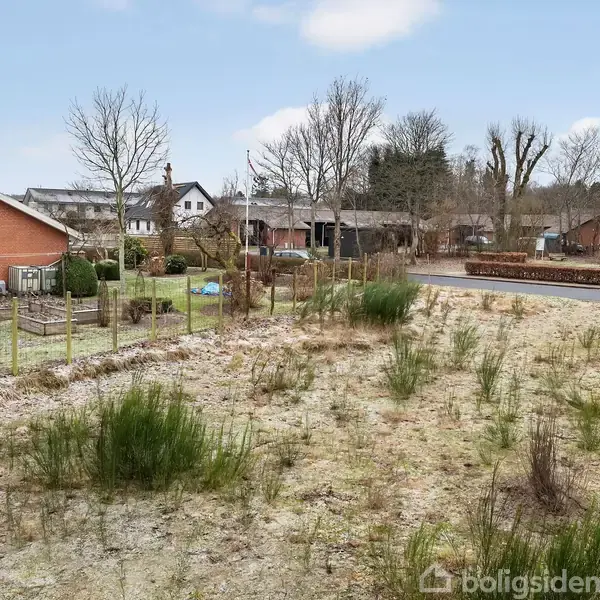 En grusbelagt jordlod med enkelte buske grænser op til et hegn mod en have. I baggrunden ses et rødt murstenshus langs en vej omgivet af træer.