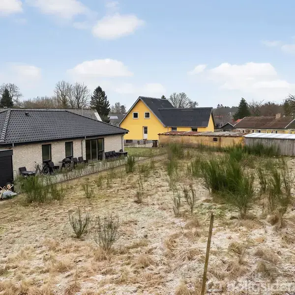 Et ubrugt græs- og buskbevokset grundstykke omgivet af hegn. Til venstre er et hus med grå tagsten og terrassemøbler. Baggrunden viser flere huse og træer.