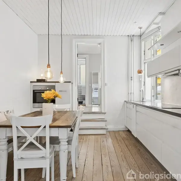 Et lyst køkken med træbord og hvide stole, prydet med en vase solblomster. Indbyggede skabe og ovn. Træfarvet gulv og ophængte lamper skaber en hyggelig atmosfære.