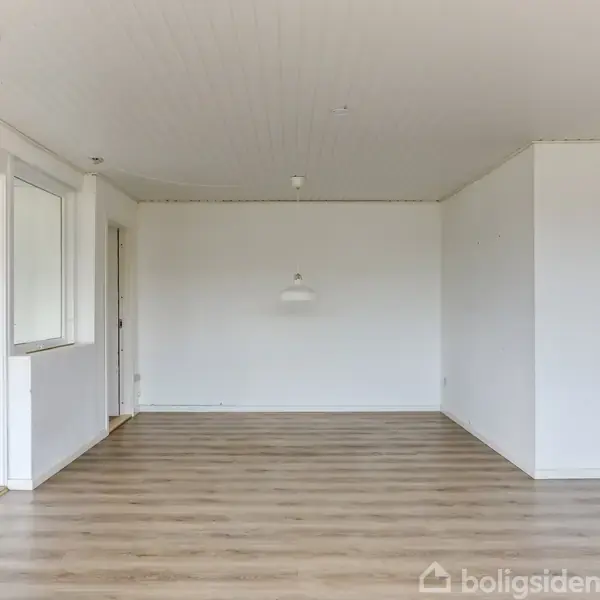 Tomt rum med lyse trægulve og hvide vægge. En enkel loftslampe hænger midt i rummet, og der findes en dør til venstre. Huller i loftet bemærkes.