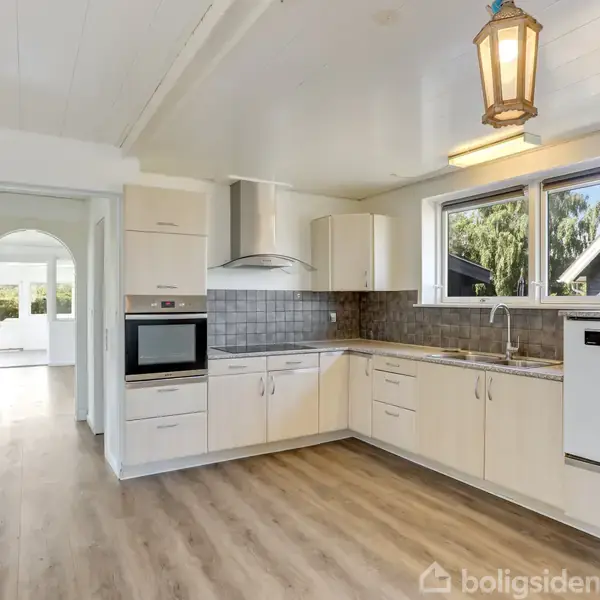 Et lyst køkken med hvide skabe og grå fliser, hvor en indbygget ovn og opvaskemaskine er synlige. I baggrunden ses en åben døråbning til en lys stue.