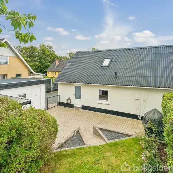 Hus med sort tag står stille i en grøn have med velplejede buske omkring. Flisebelagt terrasse foran huset. Solrig dag med blå himmel og få skyer.