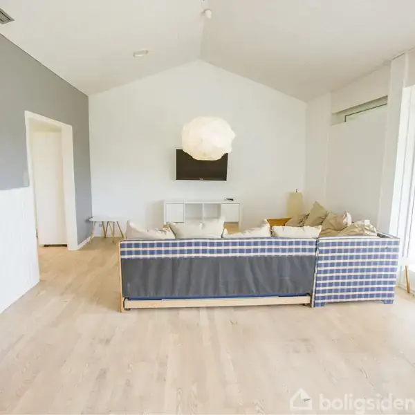 En stor, vinklet sofa står i et lyst stueområde med trægulv og hvidmalede vægge. Over sofaen hænger en stor, rund lampe. I baggrunden er en fladskærm på væggen.