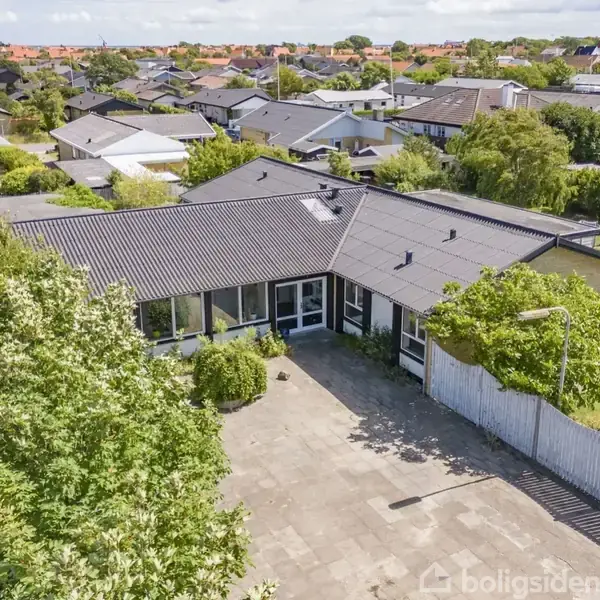 Et enkelt-etagers hus med mørkt tag og store vinduer. Huset ligger i et villakvarter omgivet af træer og en flisebelagt gårdsplads. Andre huse ses i baggrunden.