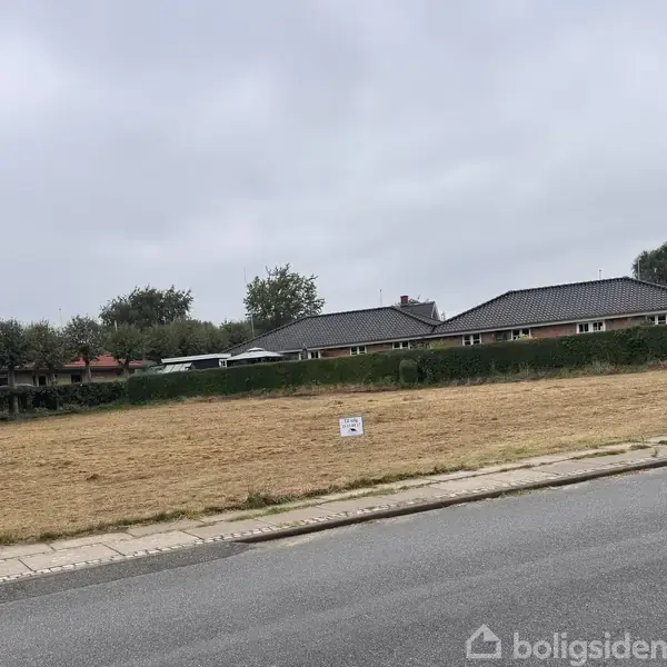 Græsplæne uden bygninger, beliggende i boligområde med rækkehuse i baggrunden. Et Lille skilt står midt på plænen. Gaden løber langs forgrunden. Skyet himmel ovenover.