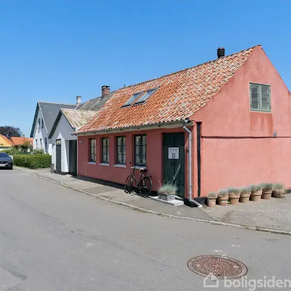 En lyserød husbygning med skråt tag står stille i en gade omgivet af andre huse. En cykel står op ad husmuren. Der er potter opstillet ved indgangen.