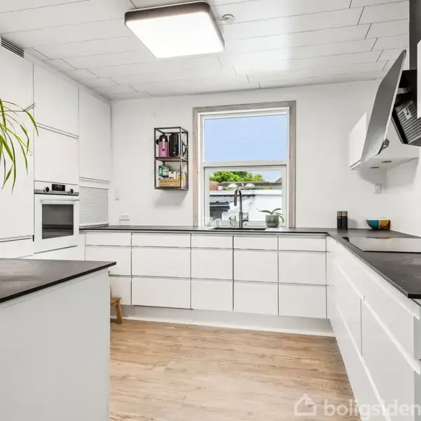 Køkken med hvide skabe og sorte bordplader, tilberedt med planter på hylderne. Vindue mod udendørs have, hvilket skaber lys og rummelig atmosfære. Trægulve og moderne hvidevarer fuldender rummet.