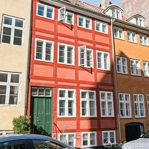 Et rødt bindingsværkshus med åbne vinduer står midt mellem to andre farvestrålende bygninger. Grøn hoveddør med "17" til venstre. Sollys skaber skygger på facaden.